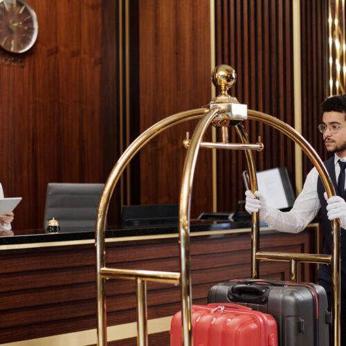 Young bellboy in uniform pushing cart with suitcases while walking along reception counter with female manager using digital tablet
